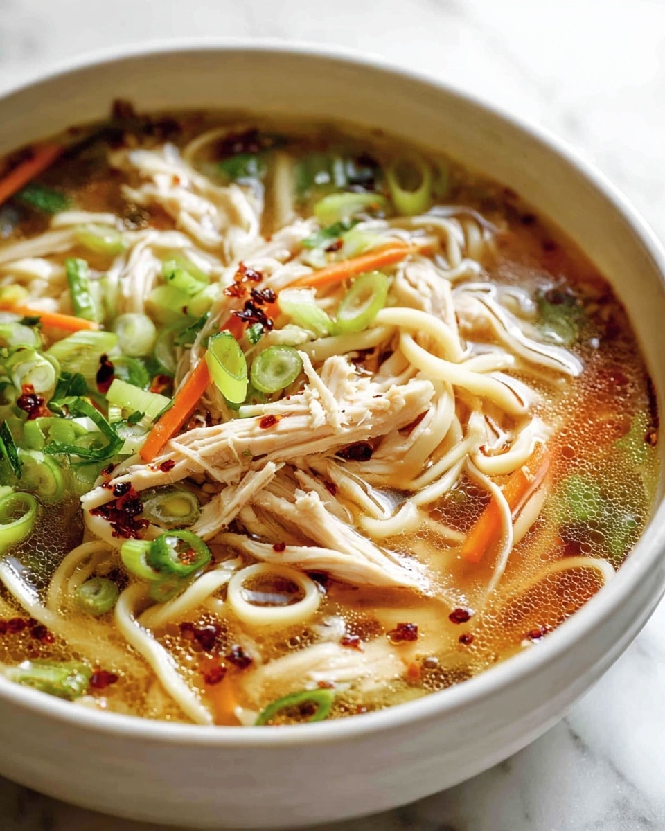 Large white bowl filled with a steaming hot serving of homemade chicken noodle soup, showcasing long, tender egg noodles intertwined with shredded chicken, sliced green onions, thin carrot sticks, and a clear, flavorful broth sprinkled with chili flakes and herbs, the entire bowl presented on a white marble surface with natural lighting, photographed from a close 3/4 angle to emphasize warmth and comfort, professional food styling photo taken with an iphone --ar 4:5 --v 7