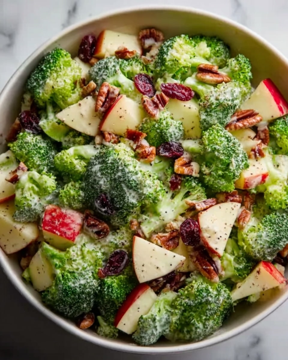 Large white bowl filled with a vibrant broccoli salad featuring crisp broccoli florets, evenly mixed with juicy diced red apples and crunchy toasted pecans, all lightly coated in a creamy dressing with visible specks of black pepper, garnished with scattered dried cranberries, presented on a white marble surface under natural light, professional food magazine style shot from a 3/4 angle showing the entire dish, photo taken with an iphone --ar 4:5 --v 7