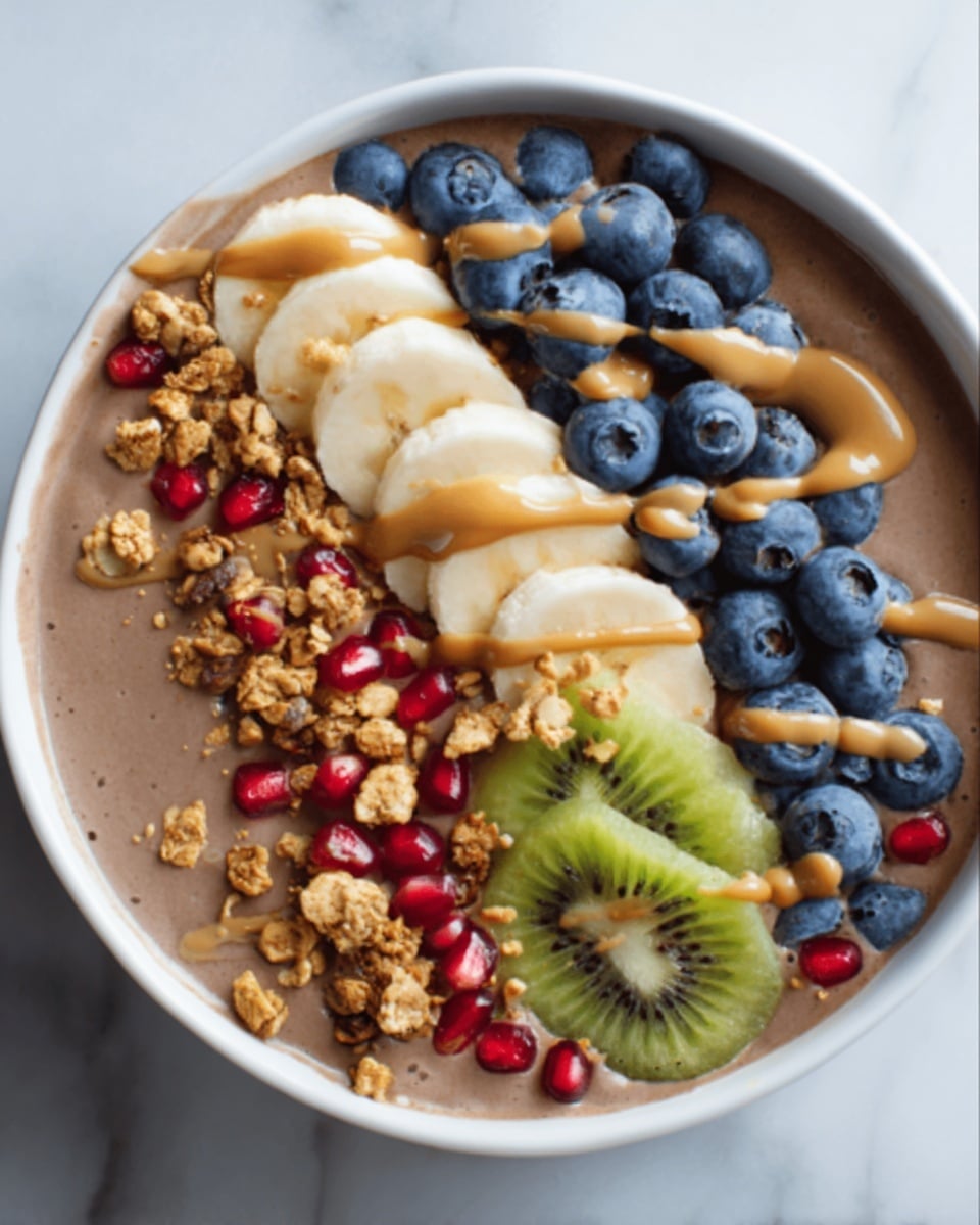 Large white bowl filled with a smooth, creamy chocolate smoothie bowl base, topped with an abundant row of fresh vibrant blueberries, thinly sliced kiwi rounds, several banana slices, crunchy golden granola clusters, and bright red pomegranate seeds neatly arranged in parallel lines across the surface, garnished with a generous drizzle of light brown nut butter, photographed from a professional 3/4 angle on a white marble countertop with natural lighting, full bowl shown in frame like a hero shot from a food magazine, photo taken with an iphone --ar 4:5 --v 7
