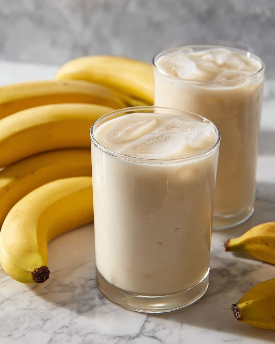 Two large white glasses filled with creamy banana smoothie with ice cubes, surrounded by fresh whole bananas arranged neatly on a white marble countertop, natural lighting highlighting the smooth texture and fresh ingredients, professional 3/4 angle food styling photo taken with an iphone --ar 4:5 --v 7