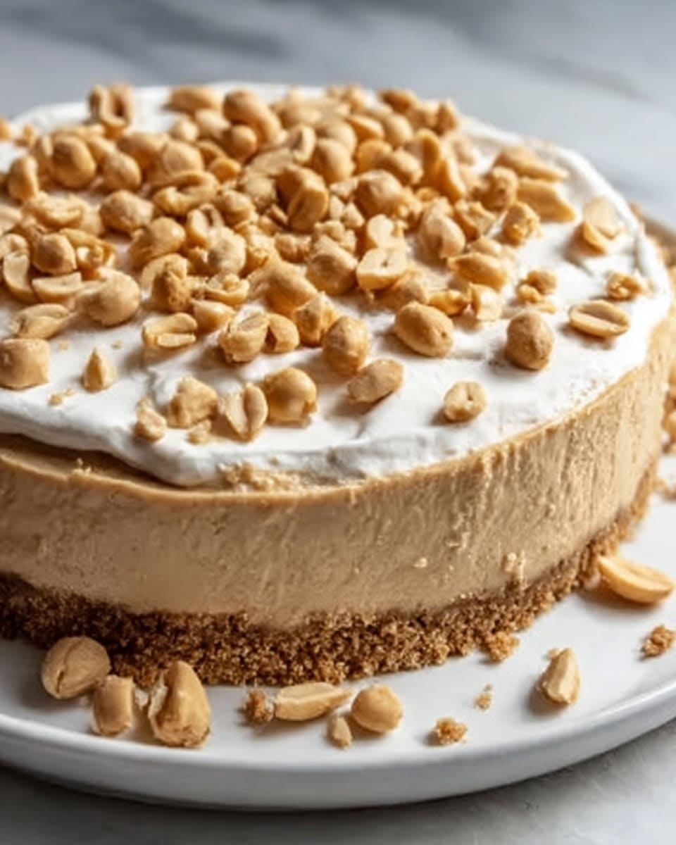 A whole, uncut peanut butter cheesecake presented in a smooth, creamy finish with a perfectly even layer of whipped topping spread over the surface, sprinkled generously with whole and halved roasted peanuts. The cheesecake sits on a thick, crumbly nut and graham cracker crust, all displayed on a large white plate. The scene is captured in natural lighting on a white marble background from a professional 3/4 angle, emphasizing the rich texture and peanut garnish for a food magazine hero shot. Photo taken with an iphone --ar 4:5 --v 7