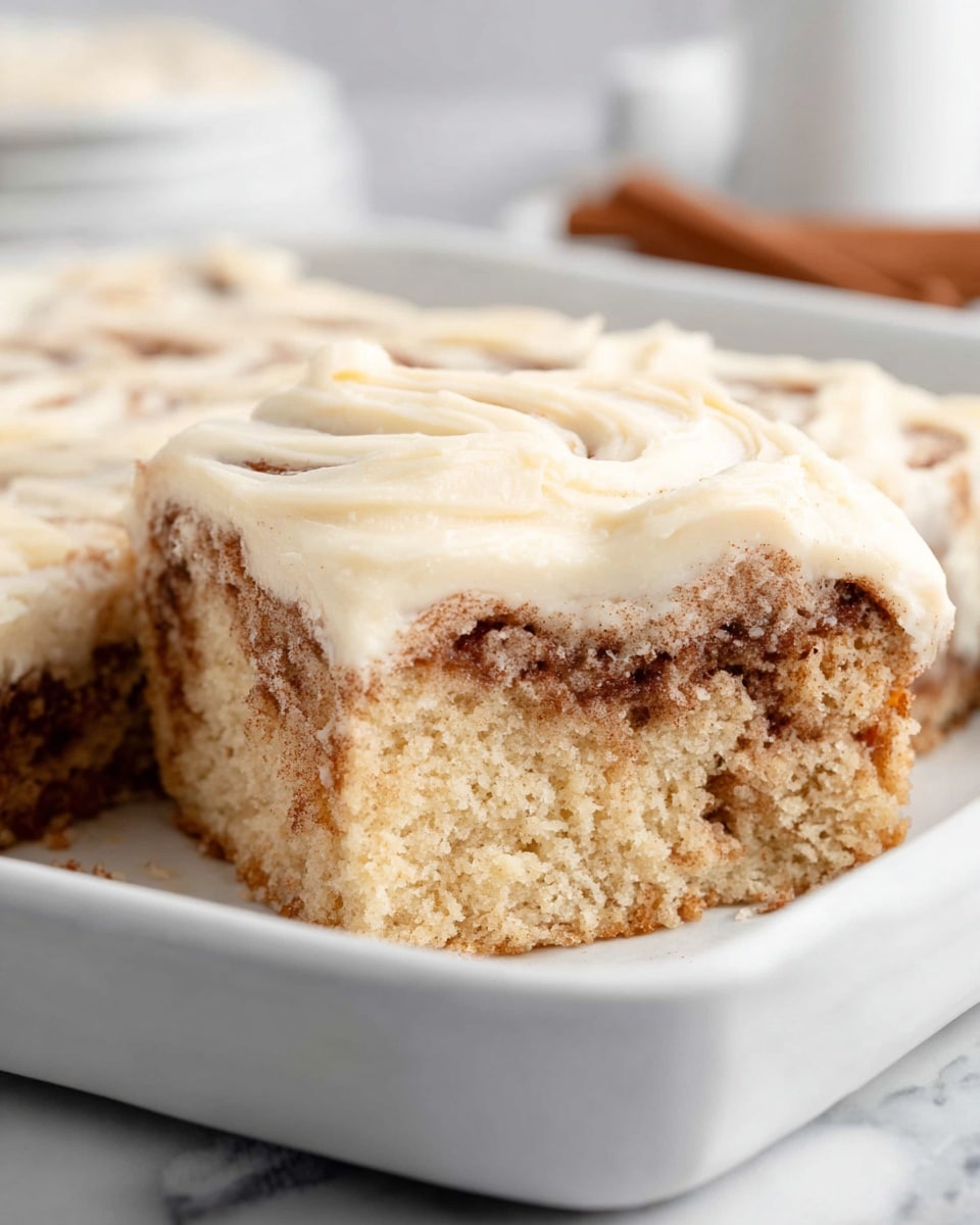 Large white rectangular baking dish filled with a fully intact cinnamon swirl cake topped with a thick layer of smooth, creamy vanilla frosting, showcasing the moist, fluffy texture of the cake and the rich cinnamon filling swirled throughout. The whole dish is captured from a professional 3/4 angle, set against a white marble countertop with natural lighting, styled as a hero shot for a food magazine. photo taken with an iphone --ar 4:5 --v 7