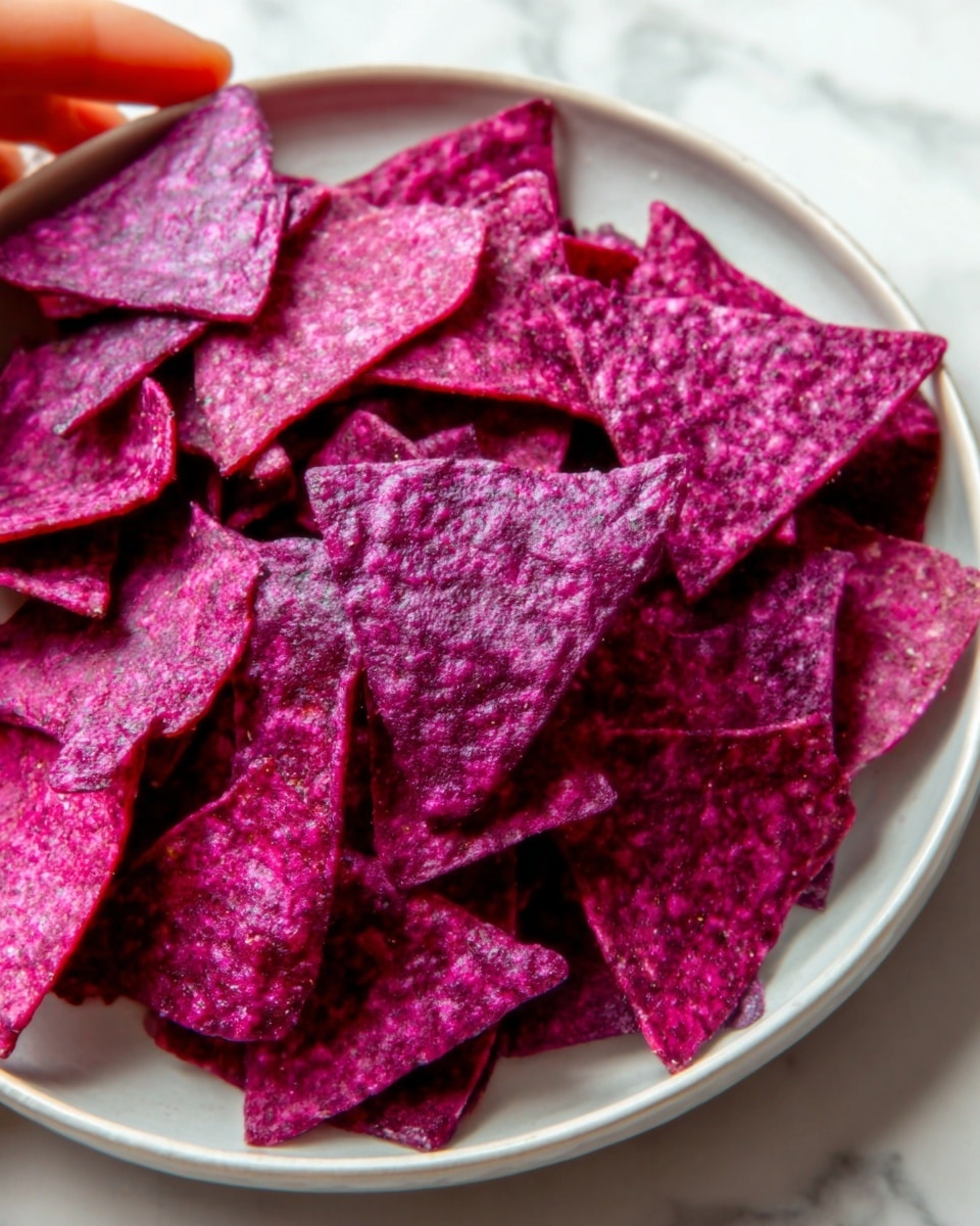 Large white bowl filled with an abundant serving of vibrant purple beet tortilla chips, showcasing their crisp, uneven edges and textured surface, all artfully piled high to emphasize the natural color and crunchiness, photographed from a slight 3/4 angle on a white marble countertop with natural lighting, styled as a striking, complete presentation like a food magazine hero shot, photo taken with an iphone --ar 4:5 --v 7