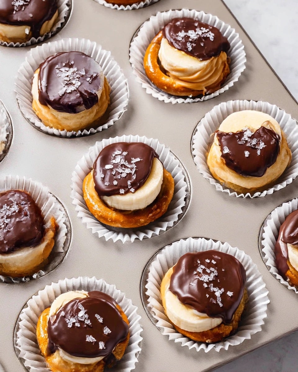 A full muffin tin lined with white paper cups, each filled with a complete peanut butter and banana pretzel bite featuring a whole pretzel base topped with a thick banana slice, a generous dollop of creamy peanut butter, and a glossy layer of rich chocolate ganache sprinkled with flaky sea salt crystals, perfectly arranged in the tray, shot at a professional 3/4 angle on white marble countertop with natural lighting, showcasing the entire delicious batch, photo taken with an iphone --ar 4:5 --v 7